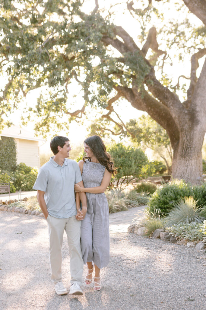 A couple joyfully walking hand in hand down a gravel path, surrounded by lush greenery and a large oak tree in the background.